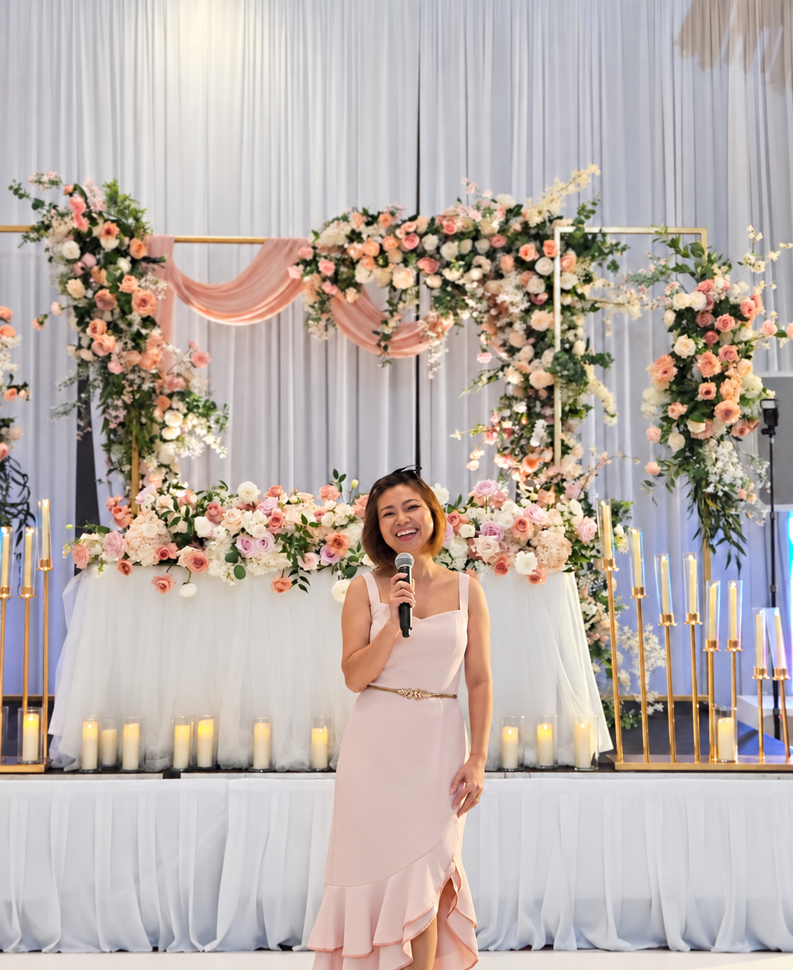 Camy Tran smiling with a microphone at a wedding, surrounded by pink and ivory flowers